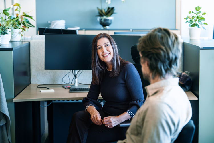 To personer sitter mot hverandre i et kontorlandskap. Vi ser bakhodet til en mann som ser på en dame vendt mot kamera. Hun smiler stort.